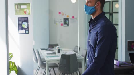 caucasian man wearing face mask crossing his arms and looking out of window at modern office