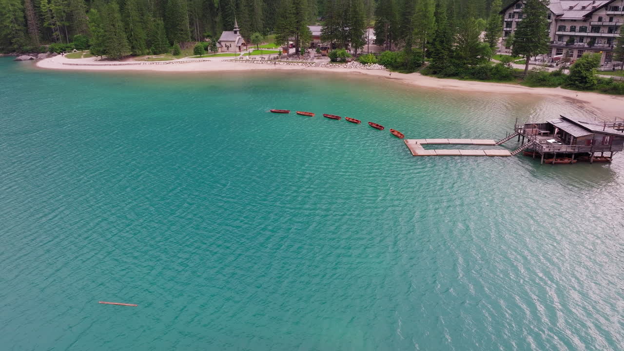 High aerial over turquoise lake in Dolomites with wooden boats and lush forest edge