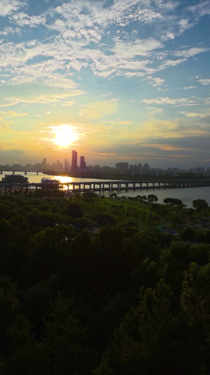 Plano aéreo vertical feito com drone com zoom no sol poente e brilhante, capturando o reflexo dourado no rio Han e no horizonte da cidade de Seul com suas pontes.