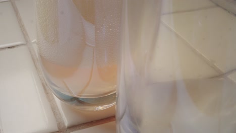 Macro-close-up-of-the-bottoms-of-stemless-wine-glasses-on-a-white-tile-kitchen-counter-being-filled-with-sparkling-ros?