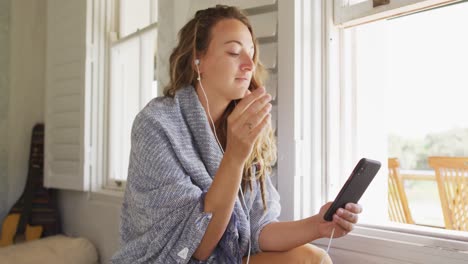 Feliz-Mujer-Caucásica-Con-Auriculares-Sentada-Usando-Un-Teléfono-Inteligente-En-La-Soleada-Sala-De-Estar-De-La-Cabaña