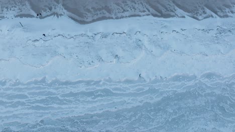 Completely-frozen-lake-shore-with-a-snowy-landscape,-serene-winter-scenery,-top-down-aerial-view