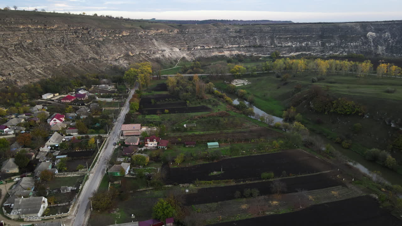 Explore a tranquil village nestled near a striking cliff and lush farms. This autumn landscape shows houses, gardens, and vibrant foliage under a clear sky