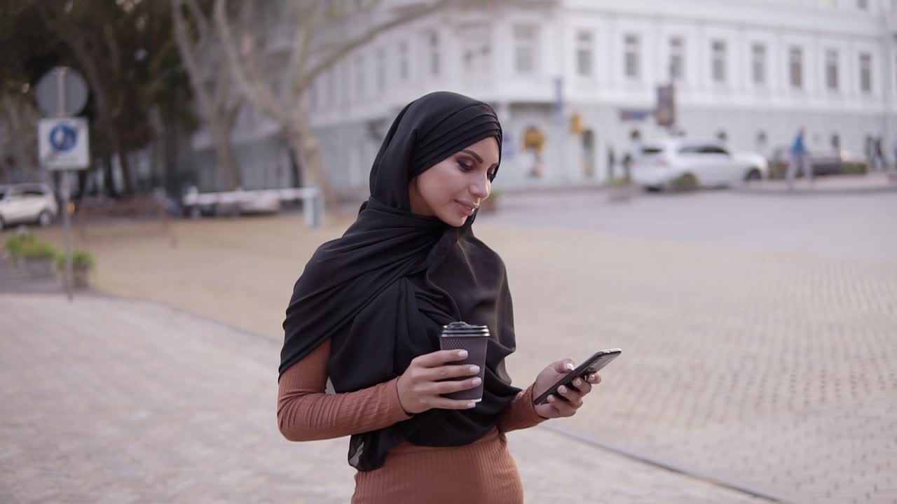 mujer joven atractiva en hiyab con un maquillaje perfecto caminando, bebiendo café y mirando la pantalla de su teléfono celular y sonriendo a la cámara