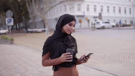 attractive young woman in hijab with a perfect make up walking, drinking coffee and looking at her cell phone screen and smiling to the camera