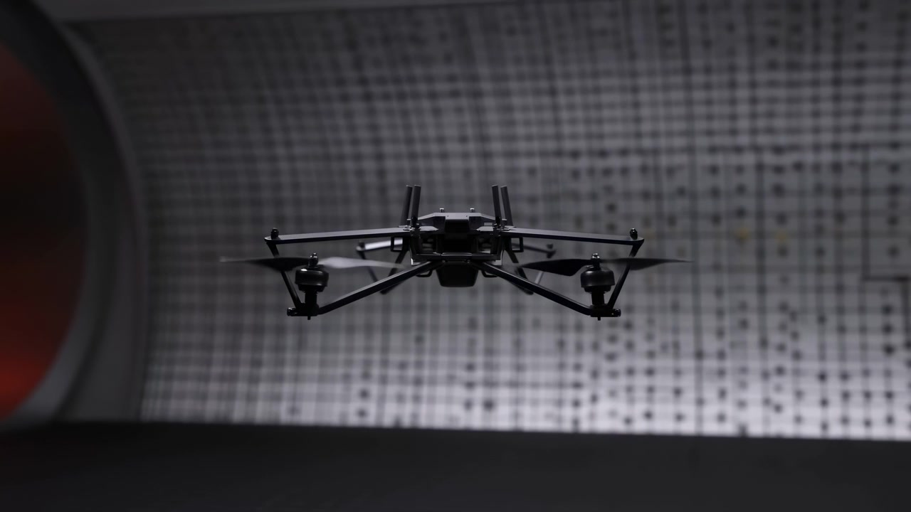 Piccolo drone rimane sospeso in aria mentre si muove leggermente attraverso l'inquadratura, con corpo nero, eliche, involucro curvo e una morbida texture a griglia illuminata che crea un'ambientazione interna futuristica