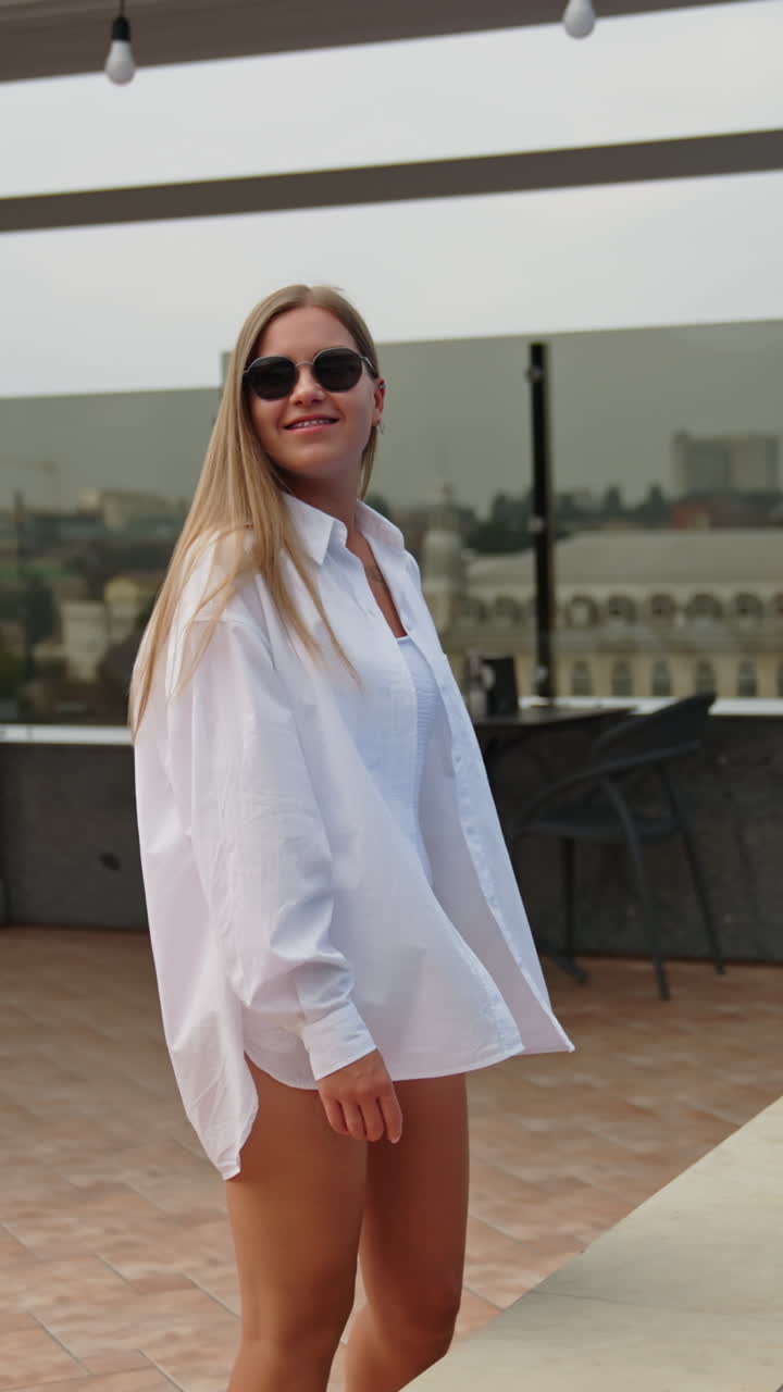 Young blonde lady in white swimsuit and shirt walks near the swimming-pool. Relaxing lounge zone at the roof of a building. Cityscape at backdrop. Vertical video