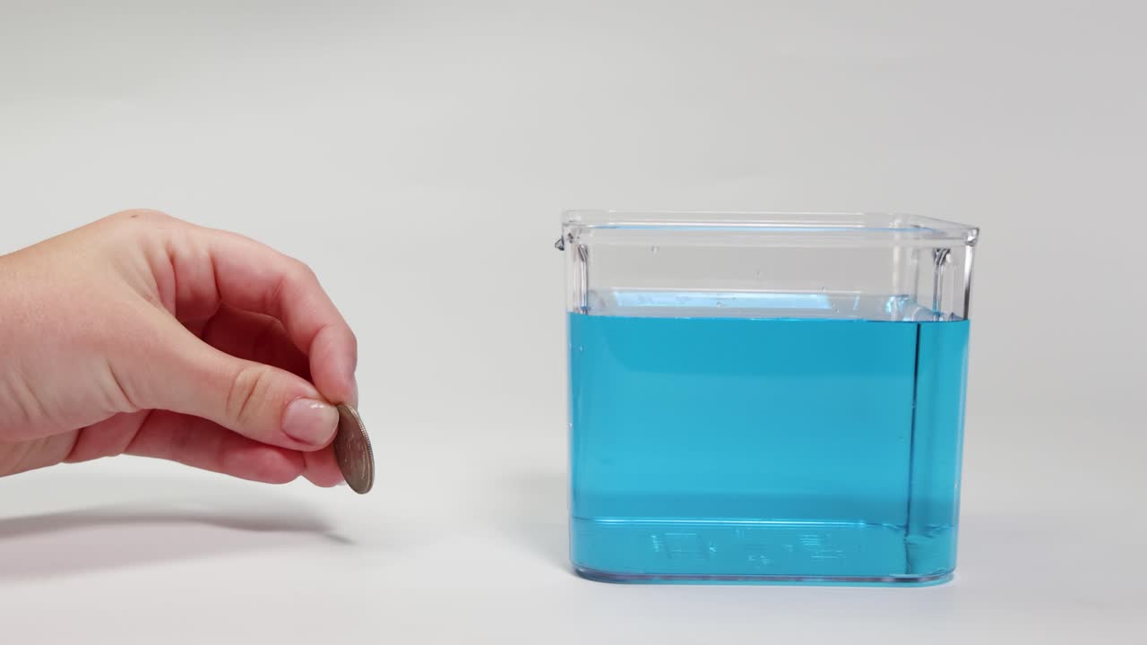 A hand drops a metal coin into a clear container filled with blue liquid, demonstrating buoyancy and density