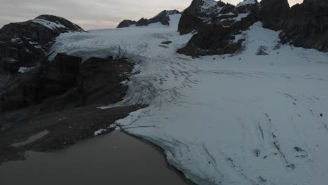 Vista-Aérea-Del-Lago-Glacial-Del-Glaciar-Claridenfirn-En-Uri,-Suiza-Al-Atardecer-Con-Una-Vista-Panorámica-Del-Cielo-Brillante-Detrás-De-Los-Picos-Alpinos-Desde-El-Agua-Llena-De-Iceberg