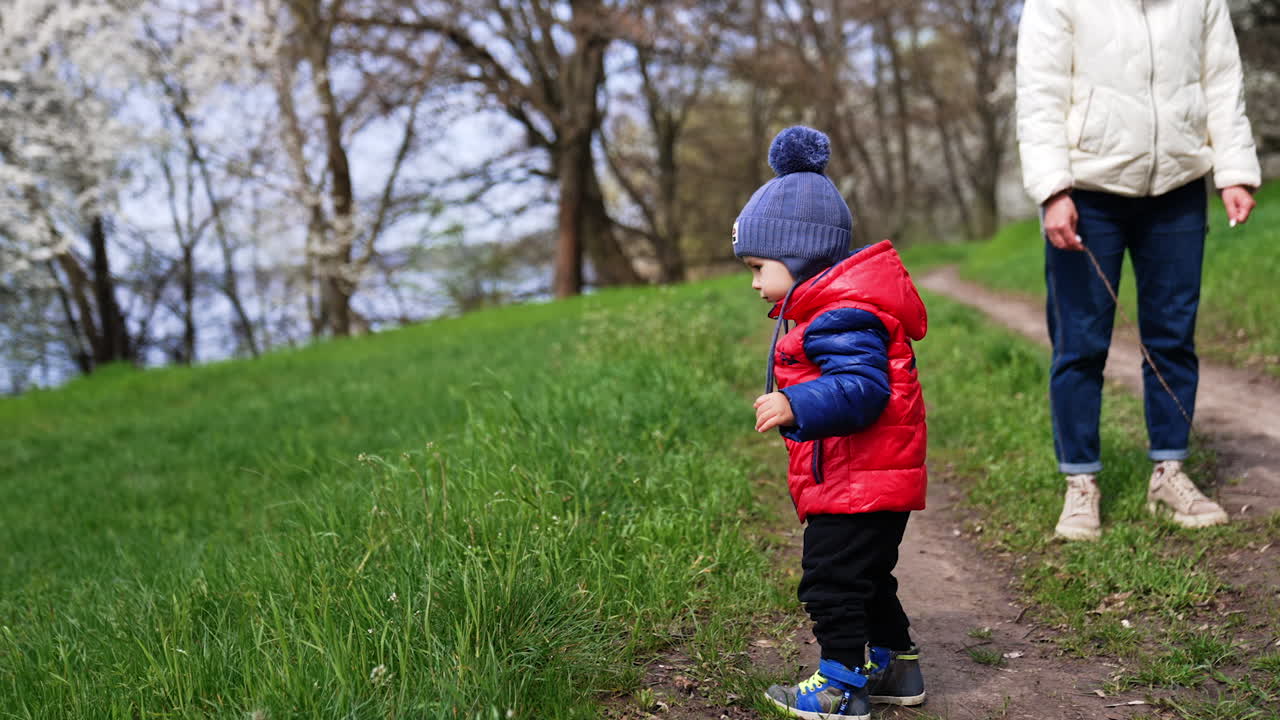 Little toddler on the walk in the nature with his mom. Baby boy in red blazer goes to green grass.