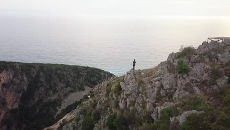 Humano-Parado-En-Un-Enorme-Acantilado-Mirando-Al-Océano-Al-Atardecer