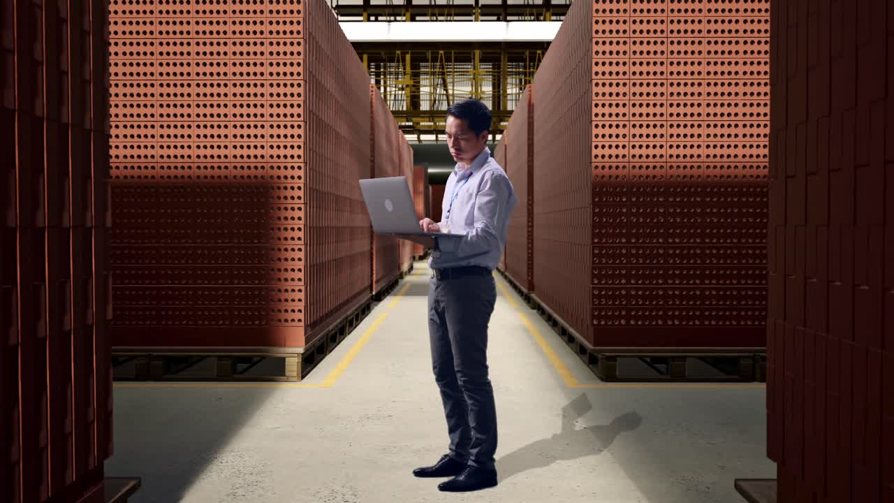 Full Body Side View Of An Asian Male Professional Worker Standing With His Laptop With Red Brick Packed in Stacks Are Stored, Typing And Dissapionted With Nodding His Head