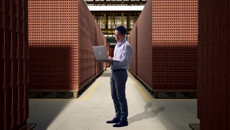 full body side view of an asian male professional worker standing with his laptop with red brick packed in stacks are stored, typing and dissapionted with nodding his head