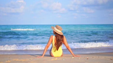 Vista-Trasera-De-La-Mujer-Sentada-En-La-Playa-De-Arena-Con-Traje-De-Baño-Amarillo-Y-Sombrero