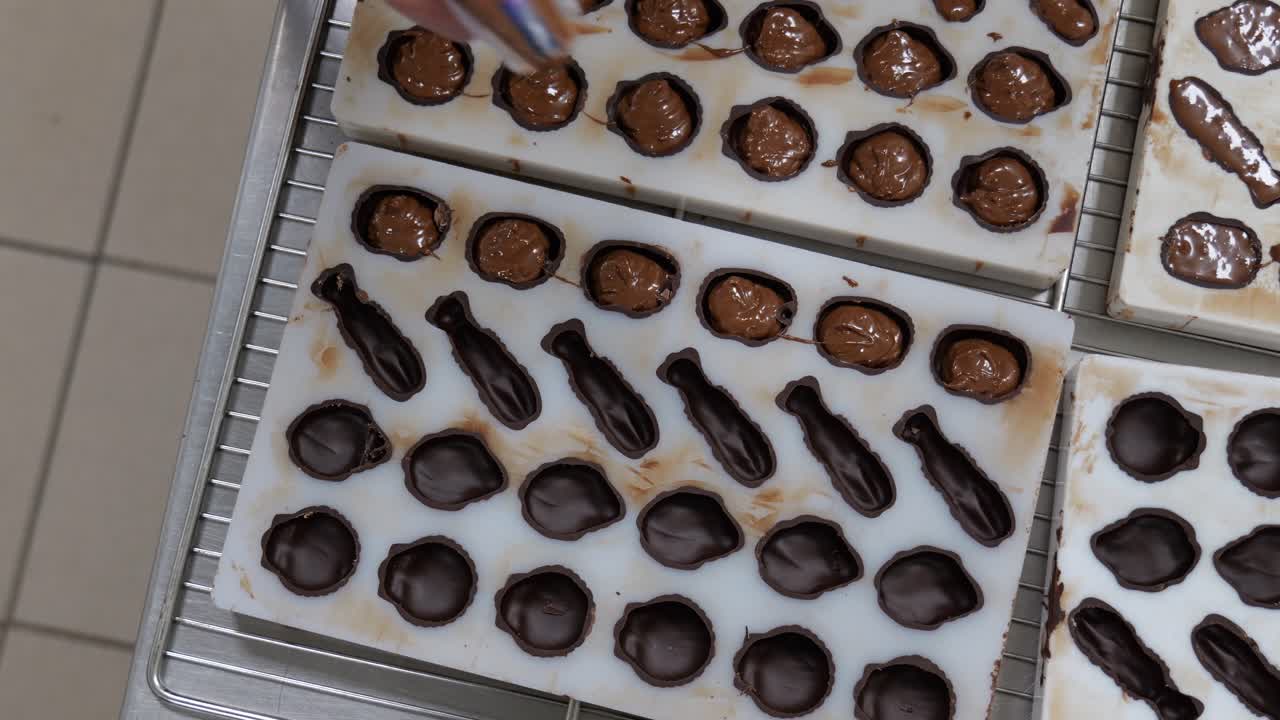 Primo piano ravvicinato dall'alto di un cioccolatiere che cola cioccolato più chiaro in stampi scuri già pronti in un laboratorio di pasticceria francese, con movimenti precisi della mano e tecnica artigianale.