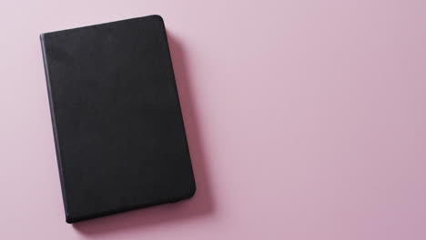 close up of closed black book with copy space on pink background in slow motion