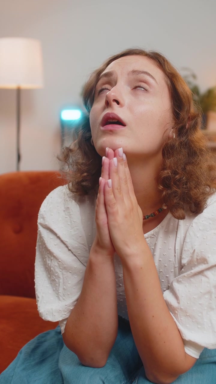Sad woman sitting on sofa at home in shock by awful news folding palms together pray ask for help