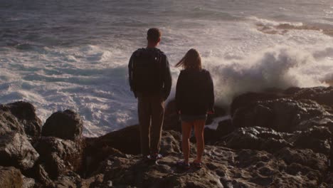 a couple stands on a rocky shore as waves roll in and crash