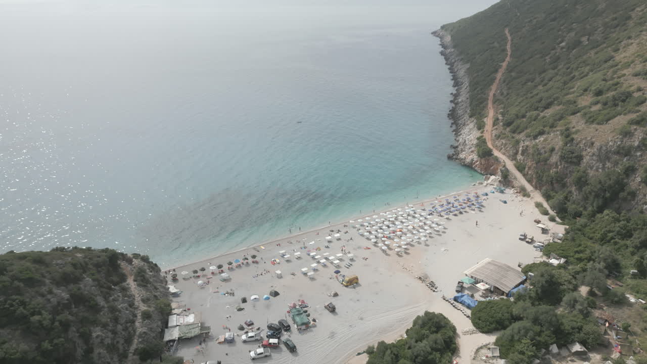 Drone shot over Gjipe beach Albania with tourists on the beach and clear blue water near the mountains and the sea on a sunny and bright day and green nature around with sun beds towels LOG