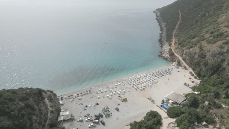 Drone-Disparó-Sobre-La-Playa-De-Gjipe-Albania-Con-Turistas-En-La-Playa-Y-Agua-Azul-Clara-Cerca-De-Las-Montañas-Y-El-Mar-En-Un-Día-Soleado-Y-Brillante-Y-Naturaleza-Verde-Alrededor-Con-Tumbonas-Y-Toallas