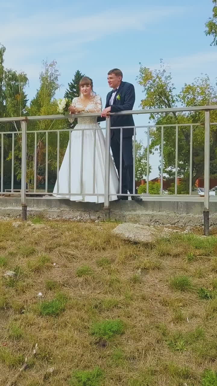 pretty girl in long wedding dress and young groom lean on metal fence railing in green park on sunny autumn day