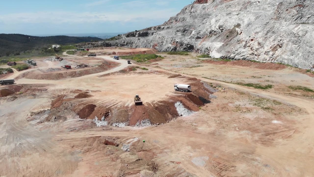 Maquinaria pesada movimenta terra em uma pedreira, cercada por formações rochosas acidentadas e solo vermelho. Adequado para projetos com temas industriais e de construção