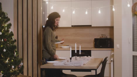 Brunette-Woman-Placing-Dishes-On-The-Table-Decorated-With-Candles-And-A-Tablecloth-For-Christmas-Dinner