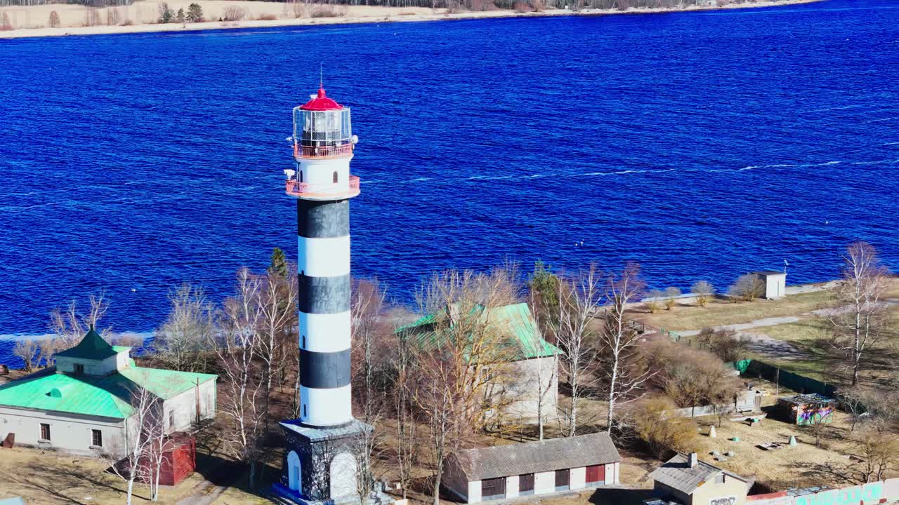A striking black and white lighthouse stands near simple houses on a rugged shoreline, framed by bare trees and deep blue water under a bright sky.