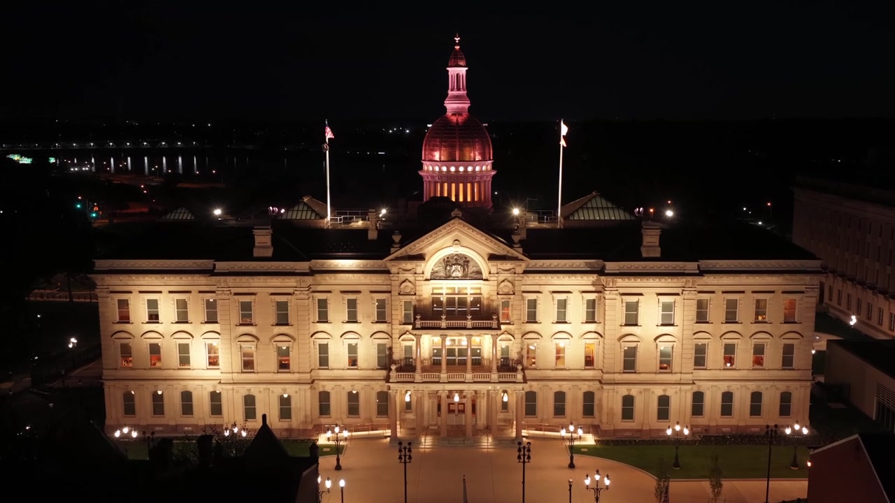 A Casa do Estado de Nova Jersey iluminada brilha à noite em Trenton, Nova Jersey, com sua cúpula de cobre ornamentada elevando-se acima da fachada histórica, iluminada por holofotes dourados perto dos mastros de bandeira abaixo. Imagem aérea ampla de drone.