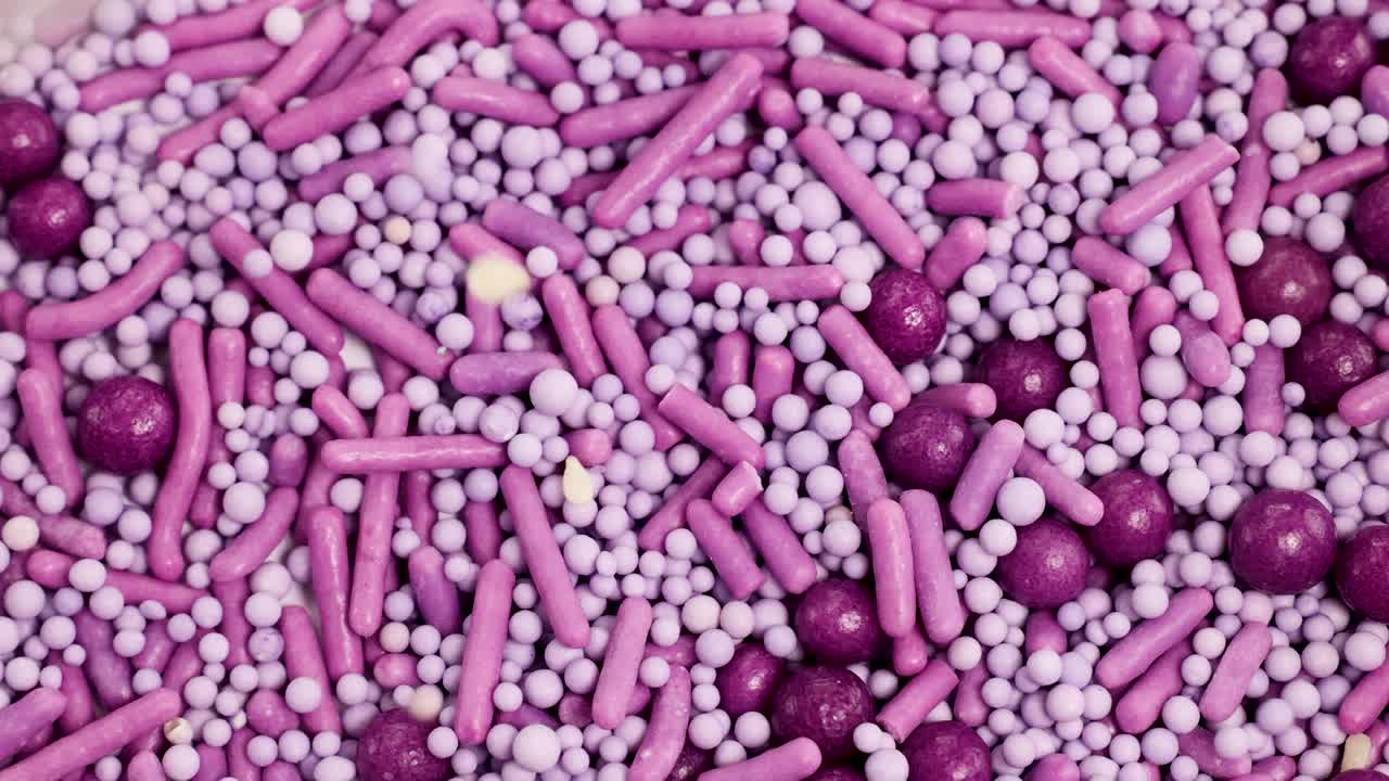 Vibrant purple and pink candy sprinkles cascade onto a surface, creating a textured, colorful pattern in macro view