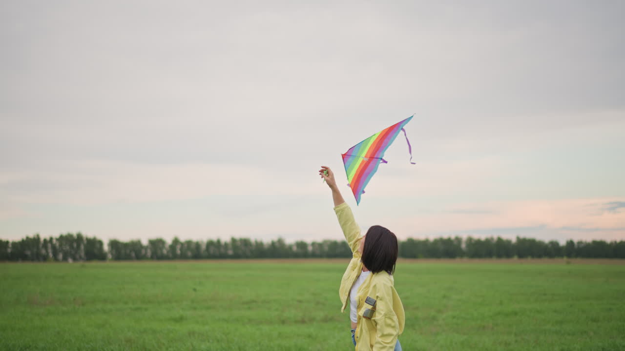 Giovane che lancia un aquilone arcobaleno in un campo, giacca gialla e jeans, in piedi da solo di fronte a un ampio orizzonte, brezza leggera che solleva la vela colorata, prato erboso e alberi in lontananza, cielo nuvoloso