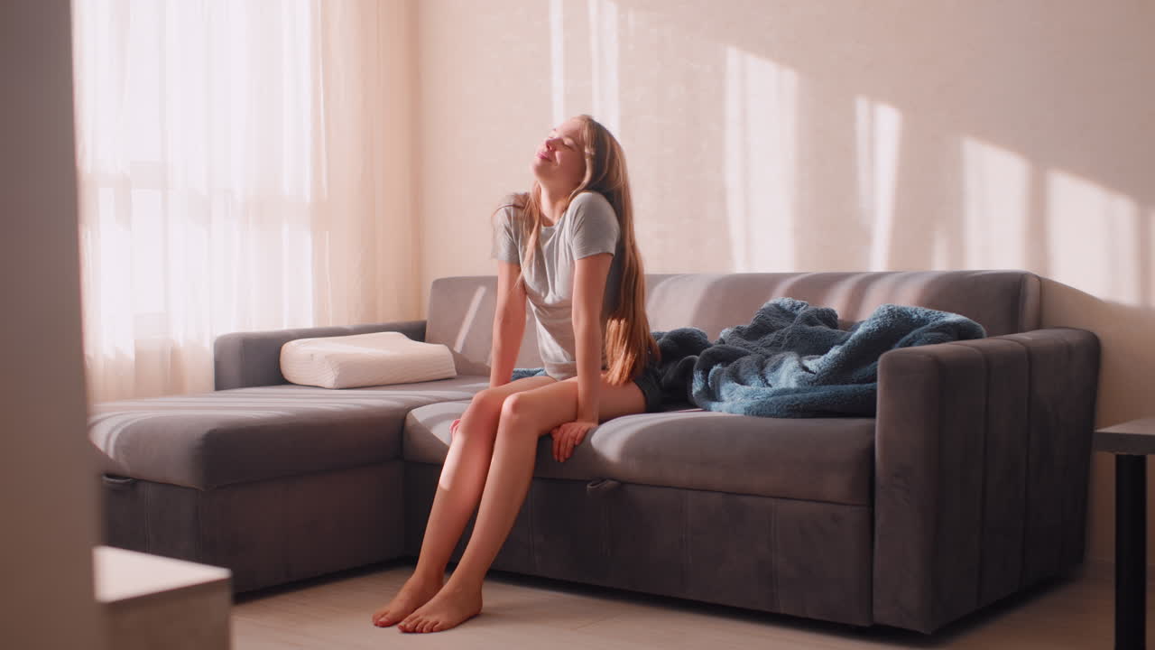 Lady seated on couch in bright room wiggles leg after waking up with smile, enjoying peaceful morning sunlight streaming through window while blanket rests beside her