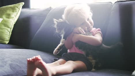 a little girl sitting on the couch holding a kitten and giving it a hug