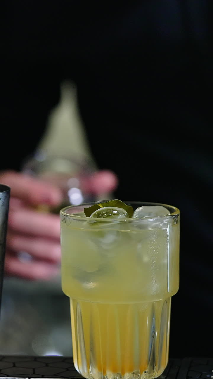Making cocktails on the bar stand. Male bartender putting a green leaf into a glass filled with ice and beverage. Close up. Blurred backdrop. Vertical video