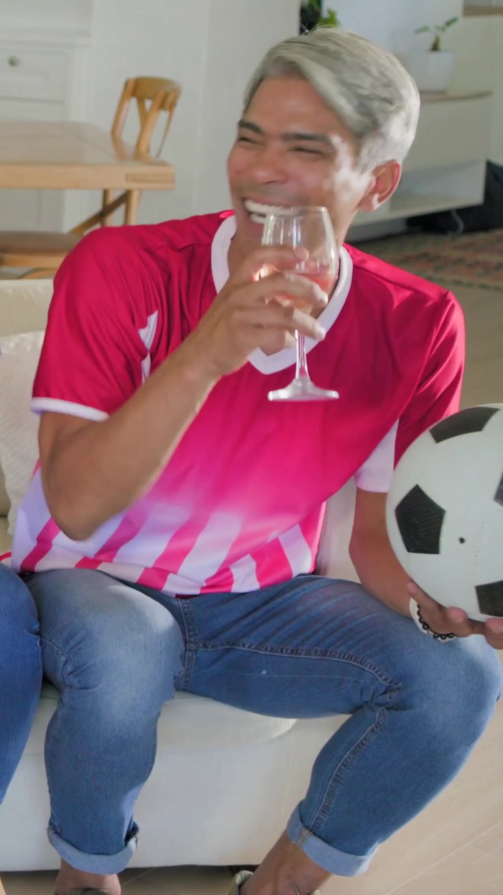 Video verticale: Uomo in maglia rossa che brinda, sorseggia vino e tiene un pallone da calcio sul divano di casa.