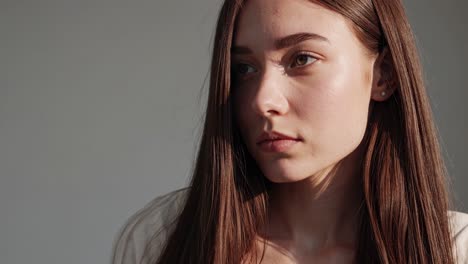 sunlight streaming through a studio, illuminating a young woman with long brown hair, enhances the contemplative mood as she poses gracefully against a soft, neutral background