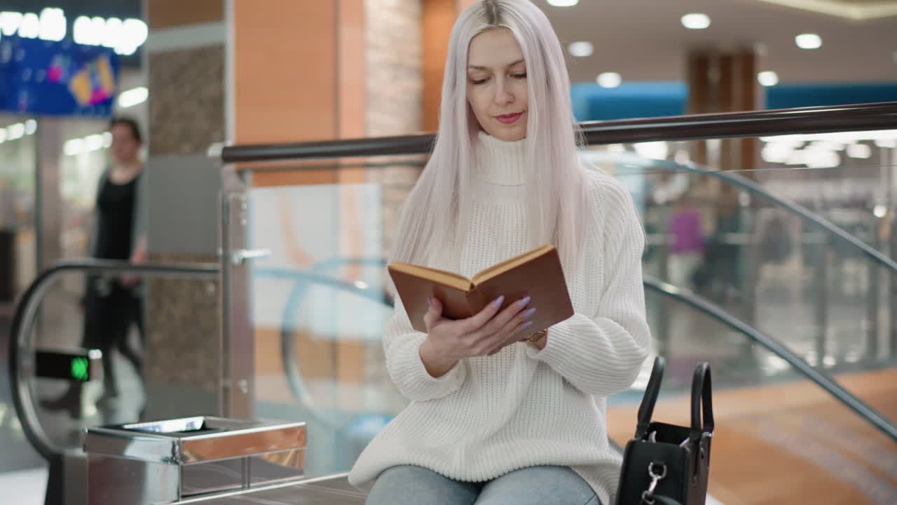 youthful lady walks to bench beside moving walkway, reaches into black bag for hardcover book, flips through pages with graceful gesture, settles into seat amid bright mall atmosphere