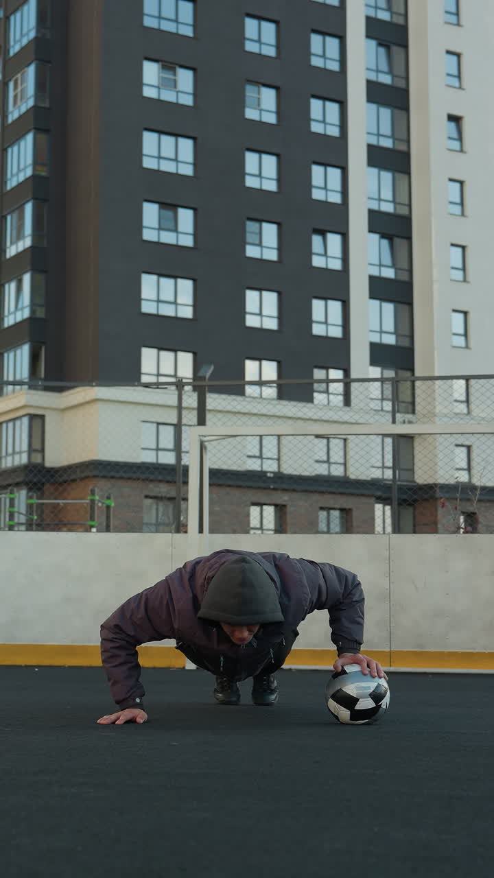 Coach performing push ups on outdoor sport arena, alternating hand placement on soccer ball, showcasing fitness with goal post and urban residential buildings in the background