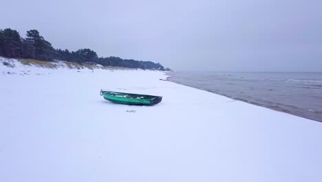 Luftaufnahme-Der-Ostseeküste-An-Einem-Bewölkten-Wintertag-Mit-Grünem-Fischerboot-An-Der-Küste,-Strand-Mit-Weißem-Sand,-Der-Von-Schnee-Bedeckt-Ist,-Küstenerosion,-Weitwinkeldrohnenaufnahme,-Die-Sich-Vorwärts-Bewegt