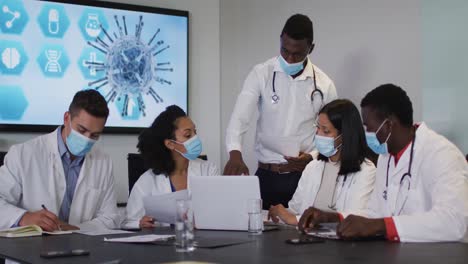 Diverse-group-of-doctors-wearing-masks-in-discussion-in-meeting-room-using-laptop