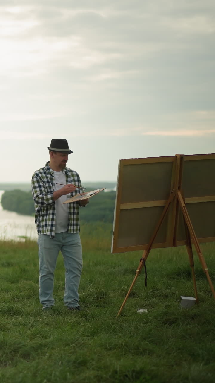 A craftsman in a black hat, checkered shirt, and jeans is engrossed in painting on a large canvas set up in a tranquil grass field by a serene lake. cloudy sky, surrounded by the beauty of nature