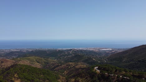 Mountains-with-winding-road-in-Spain-with-endless-ocean-in-horizon,-aerial-view