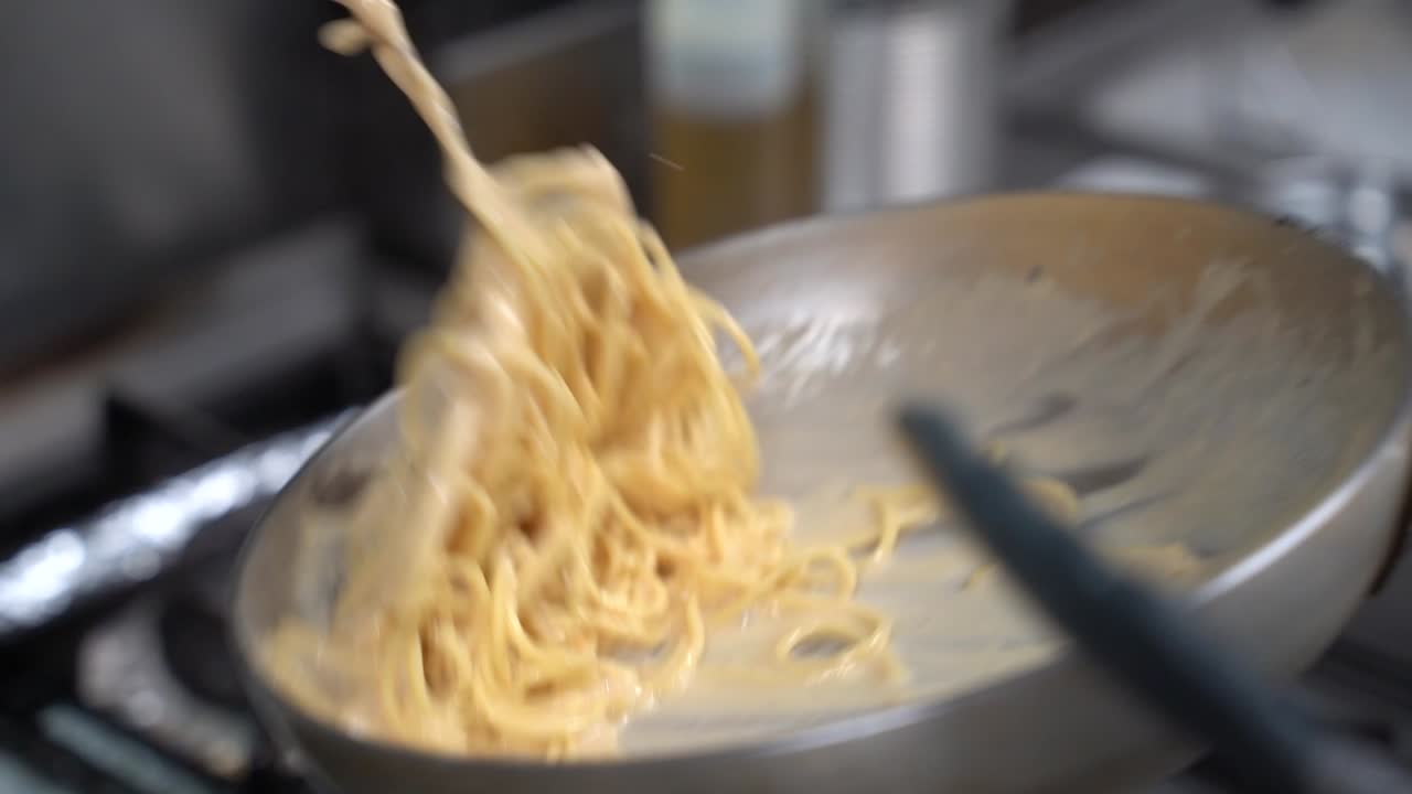 A close-up of creamy spaghetti being prepared in a large pan on a stovetop. The rich sauce coats the pasta evenly, creating a smooth and appetizing texture.