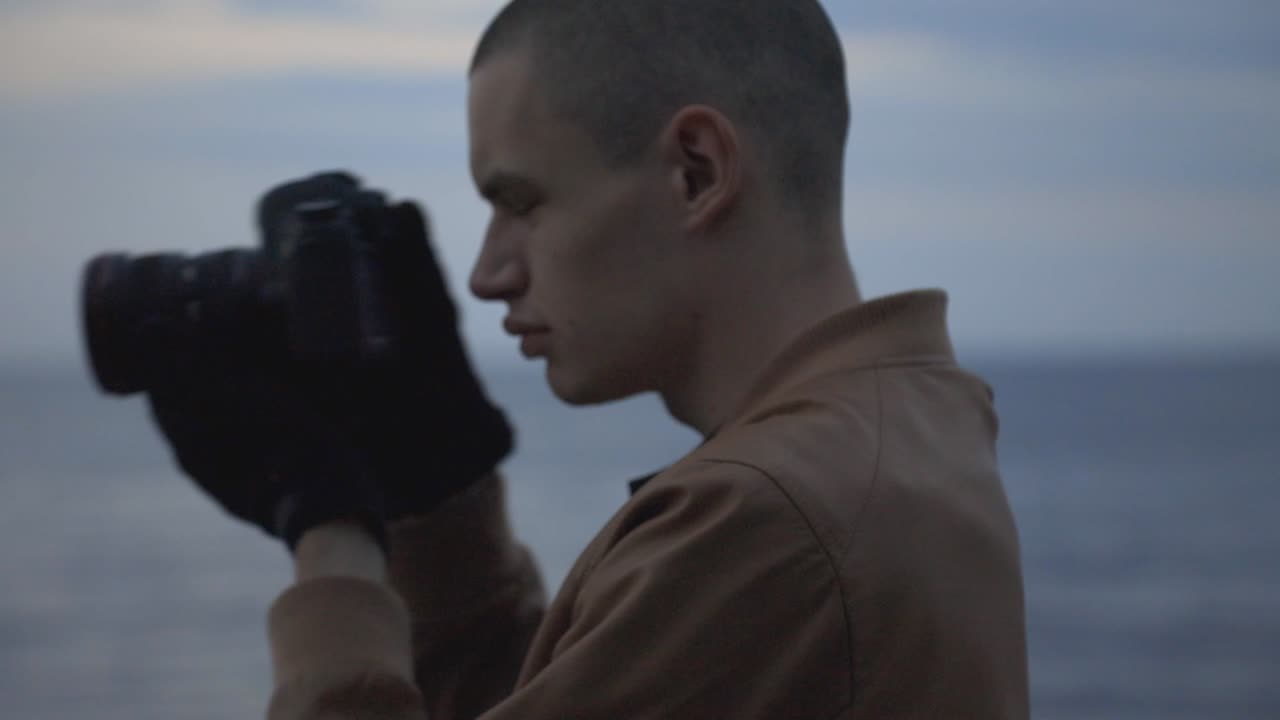 Side View Of A Young Man Standing By The Shore Wearing Black Gloves Taking Pictures With A DSLR Camera - Closeup Shot
