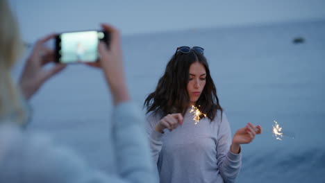 mujer bengala bailando chica usando un teléfono inteligente tomando fotos de un amigo bailando con bengalas en la playa al atardecer celebrando la víspera de año nuevo compartiendo la celebración del día de la independencia en las redes sociales