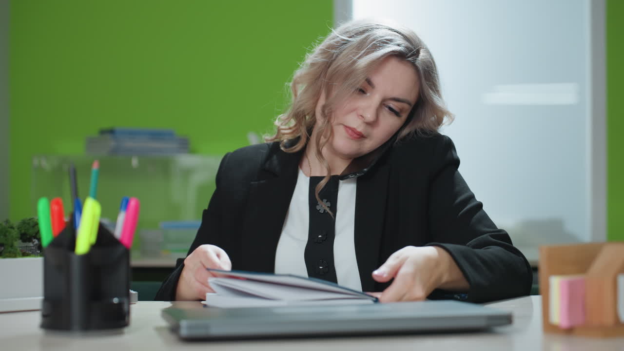 real estate agent speaks with business associate over phone, pauses with pen in hand as she prepares to record details, seated at modern office desk with architectural model and work supplies visible