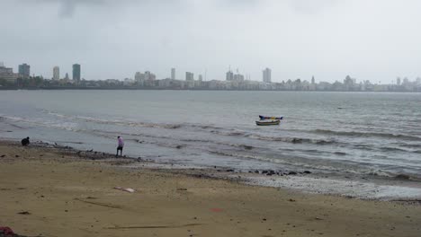 Dos-Hombres-En-La-Playa-Casi-Vacía-De-Girgaum-Chowpatty-Con-Paisaje-Urbano-En-El-Fondo-Durante-La-Pandemia-De-Coronavirus-En-Mumbai,-India