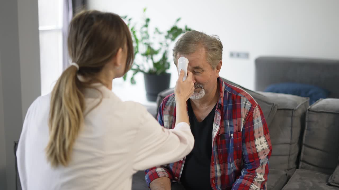 Doctor woman in lab coat takes temperature of happy senior man at home with infrared thermometer