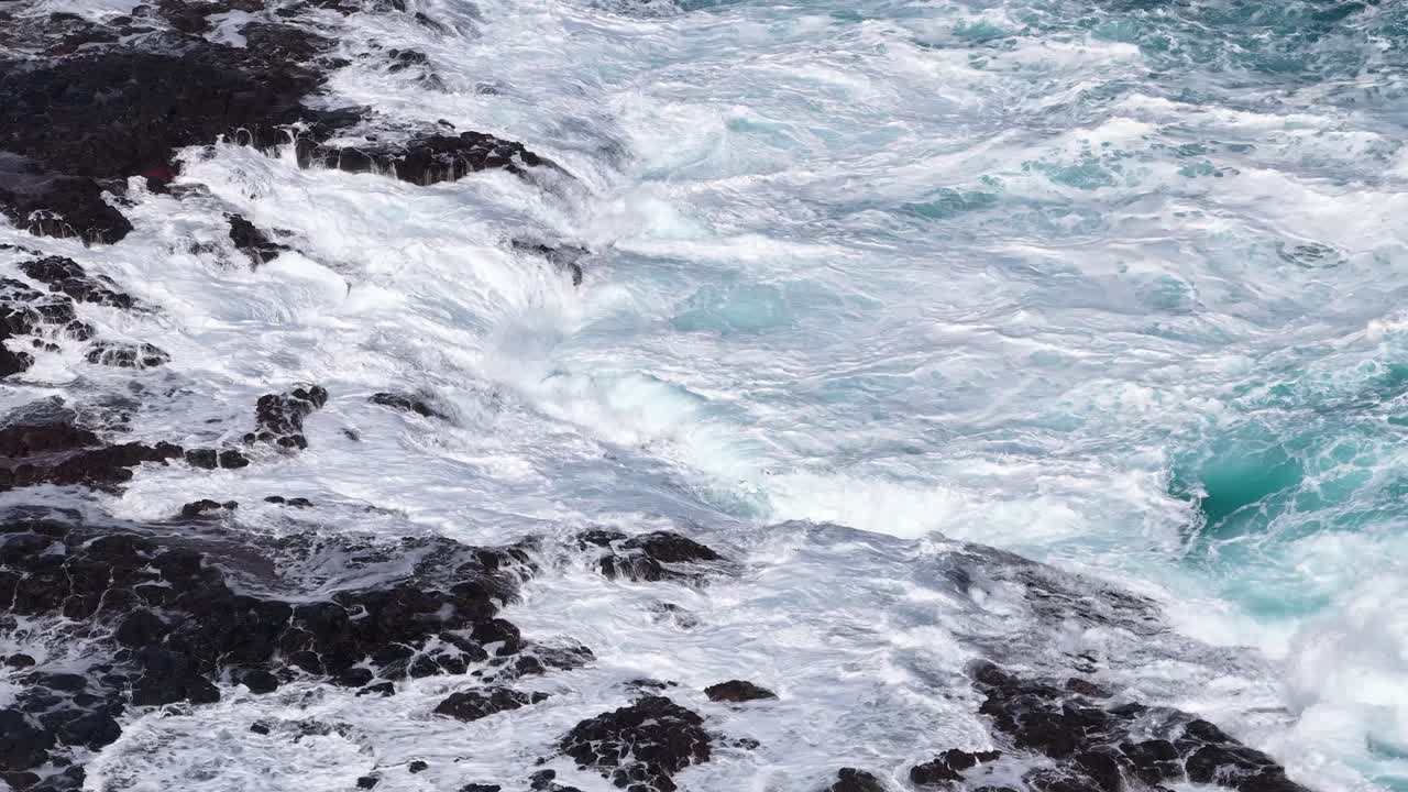Vista aerea di onde turchesi che si infrangono su rocce vulcaniche scure, luce naturale, movimento dinamico