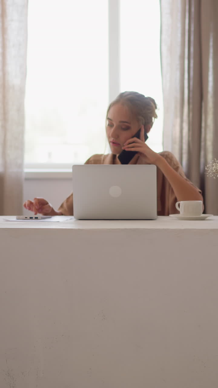 Blonde woman in stylish beige shirt works on notebook at white table talking on mobile phone with colleague against bright window in office room slow motion